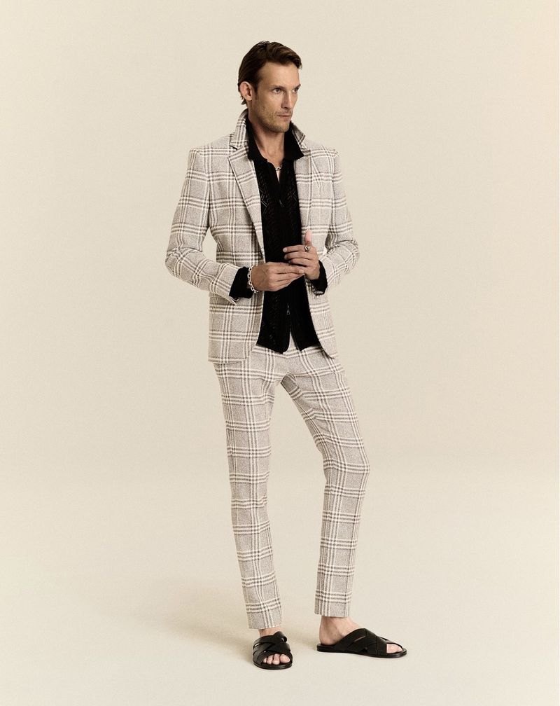 A man in a plaid suit and black shirt poses against a neutral background, wearing black leather slide sandals.
