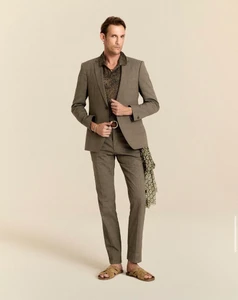 Model Pawel Binczak wearing a tailored taupe suit, patterned collared shirt, and woven slide sandals against a warm beige background.