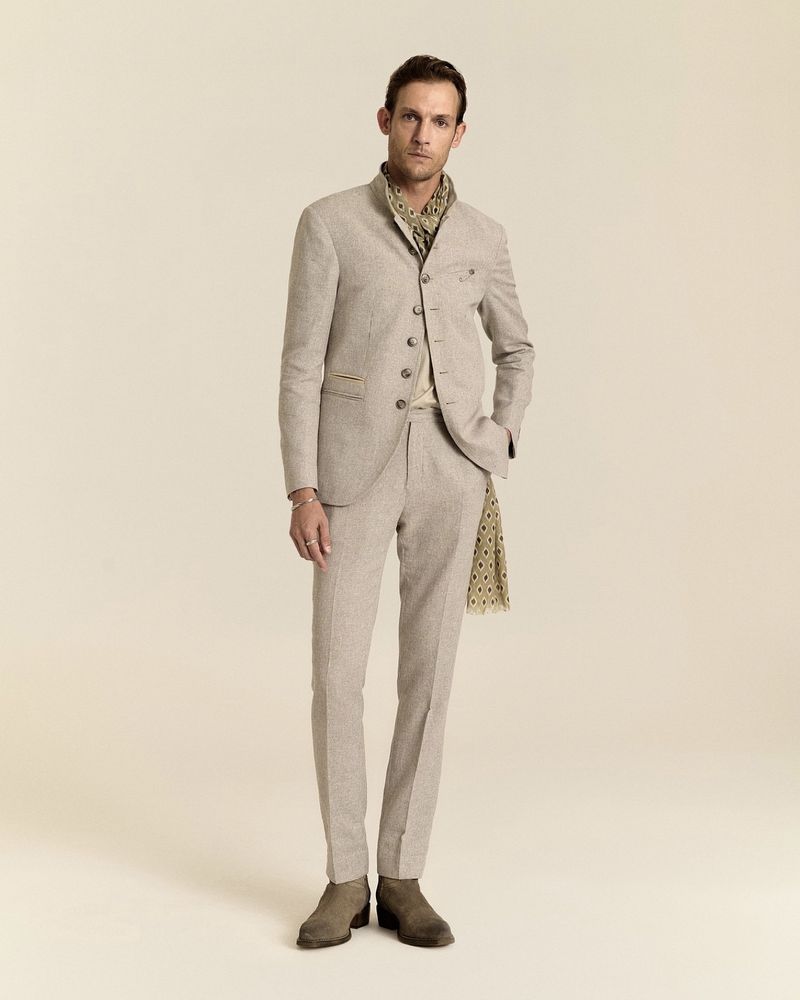 Model Pawel Binczak wearing a beige high-collar tailored suit, patterned scarf, and boots against a cream studio backdrop.