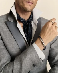Close up of a man wearing a grey suit jacket, open white shirt, silk neck scarf, and silver bracelet against a neutral backdrop.