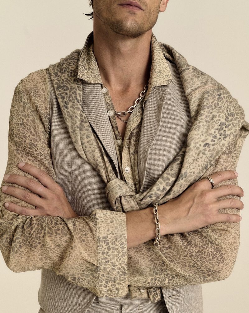 Model with arms crossed wearing a patterned button-up shirt, tailored waistcoat, tied scarf, and silver chain bracelet.