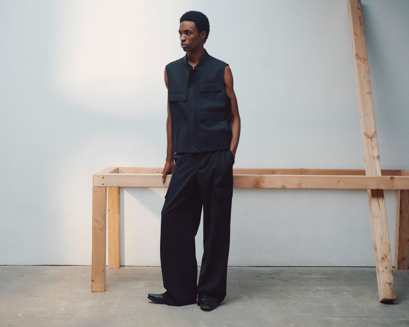 A man wearing a black utility vest and wide-leg trousers stands near a simple wooden worktable in a minimalist, light-filled space.