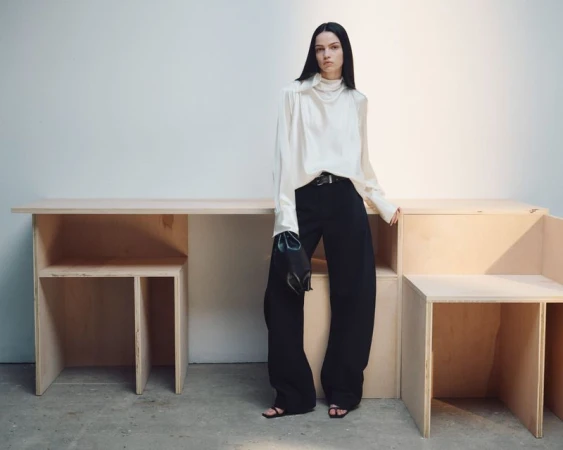 Model Sara Grace Wallerstedt in a high-neck cream blouse and wide-leg black trousers standing beside wooden furniture against a white wall.