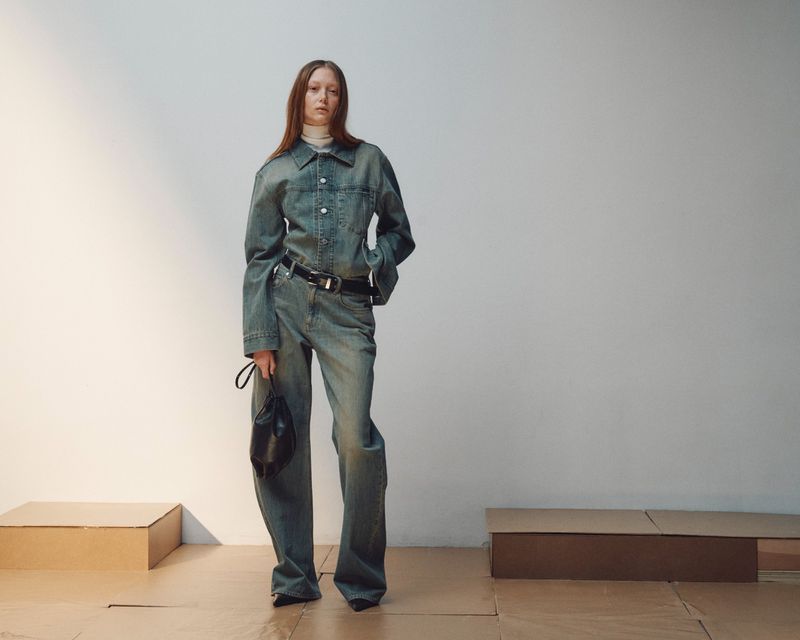 Model Sara Grace Wallerstedt wearing a matching denim shirt and wide-leg jeans while holding a black bag in a studio.