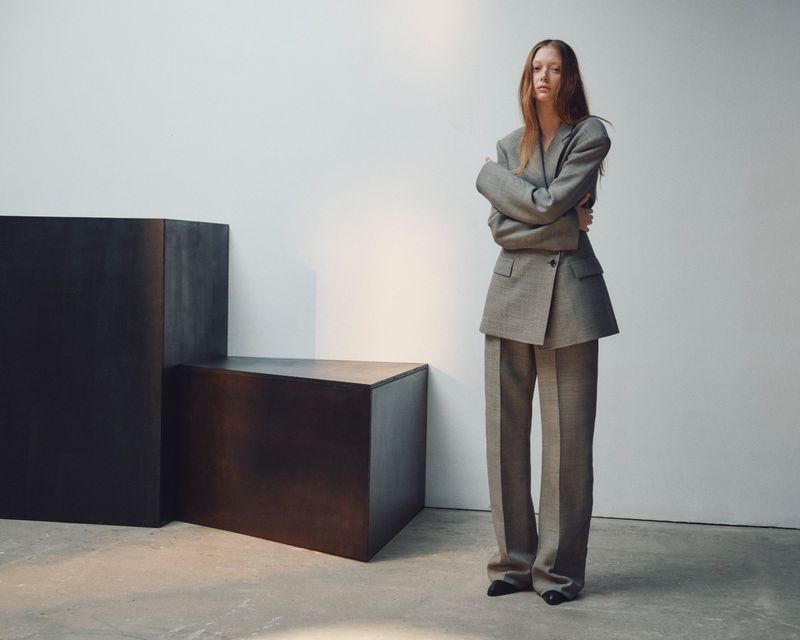 Sara Grace Wallerstedt wearing an oversized grey tailored pantsuit standing near black geometric blocks against a white wall.