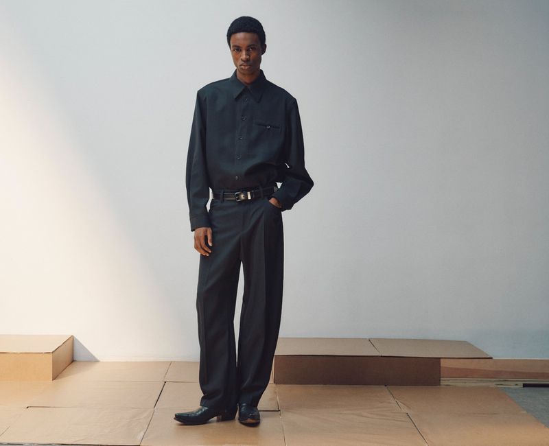 A male model wearing a dark button-up shirt and wide-leg trousers stands on cardboard staging in front of a white studio backdrop.