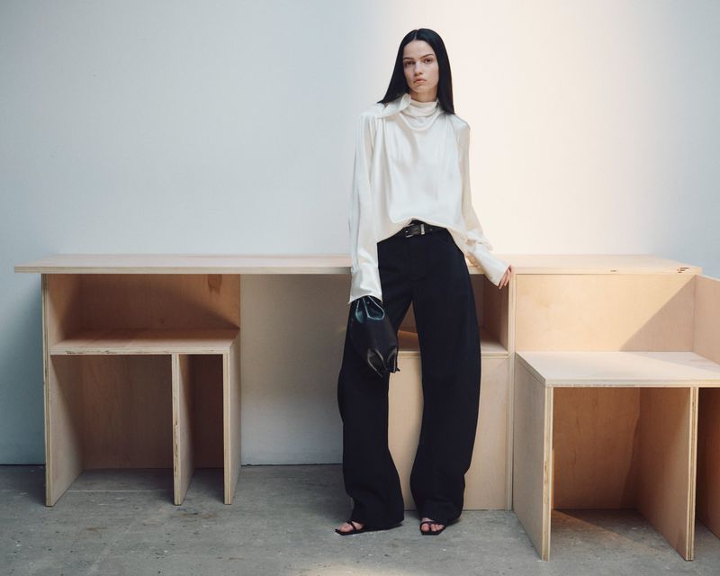Model Sara Grace Wallerstedt in a high-neck cream blouse and wide-leg black trousers standing beside wooden furniture against a white wall.