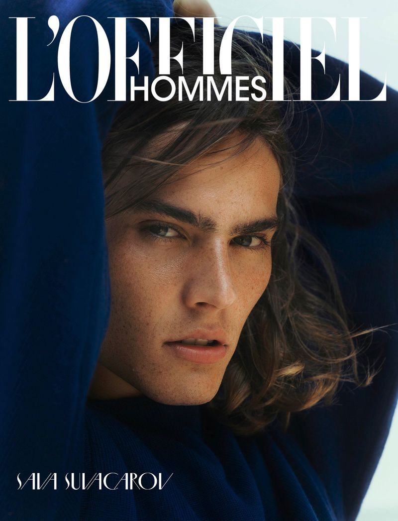 Close-up of a young man with long wavy hair and freckles wearing a deep blue hooded garment, shot for L'Officiel Hommes cover.