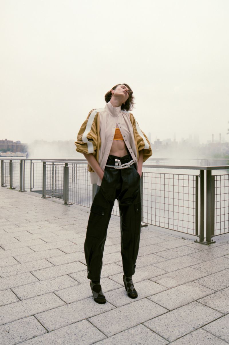 A woman in fashionable jacket, pants, and heeled shoes poses on a brick walkway with a metal railing and a cityscape in the background.