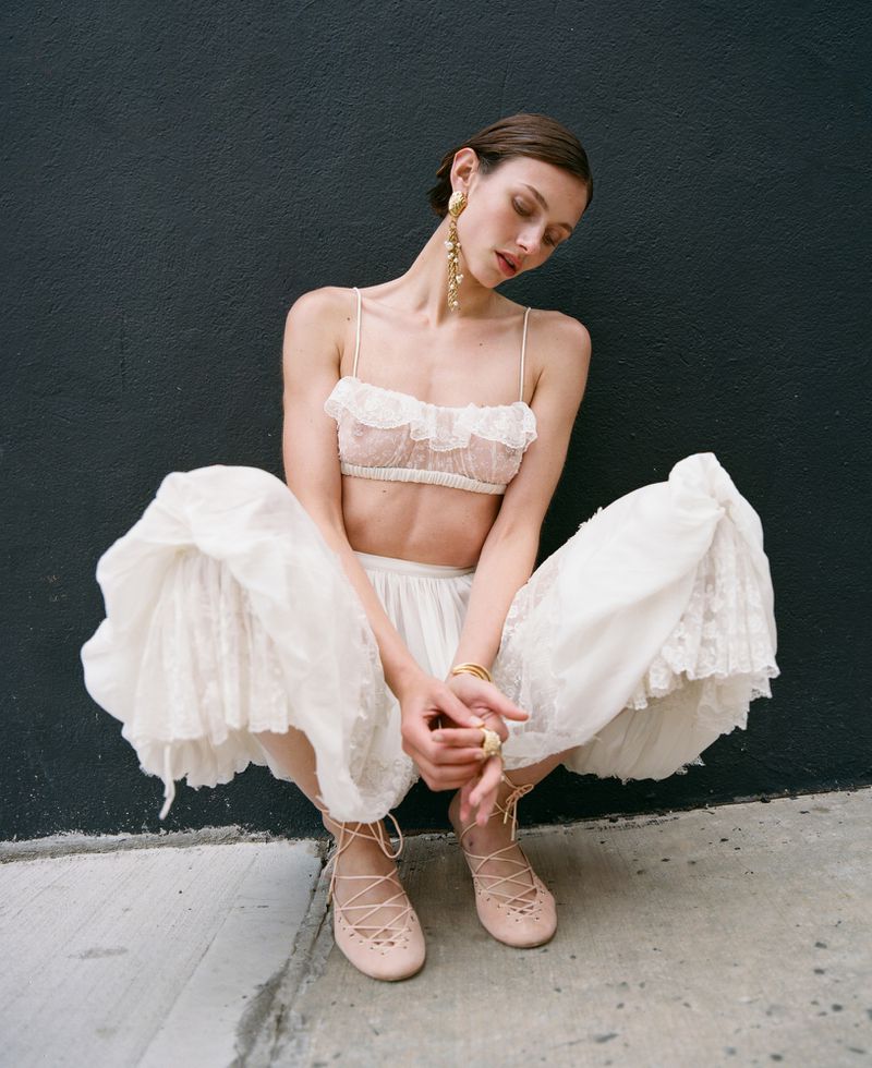 A slender woman in a white lace bralette and skirt poses against a dark wall, squatting and gazing downward in a fashion-forward outdoor setting.