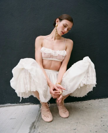 A slender woman in a white lace bralette and skirt poses against a dark wall, squatting and gazing downward in a fashion-forward outdoor setting.