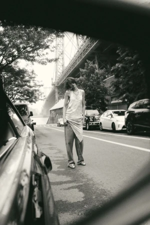 Monochrome shot of a woman in a dress and pants standing on a street, with cars parked and a large bridge in the background.