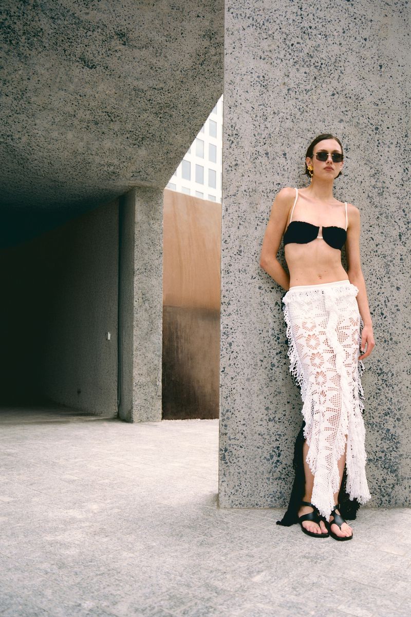 A slim adult female model poses against a concrete wall in a black bikini top and white lace skirt in an urban setting.