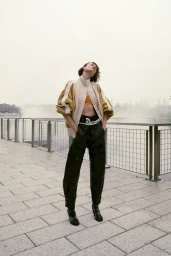 Model standing outdoors on paved waterfront promenade wearing layered jacket and dark trousers for WWD editorial
