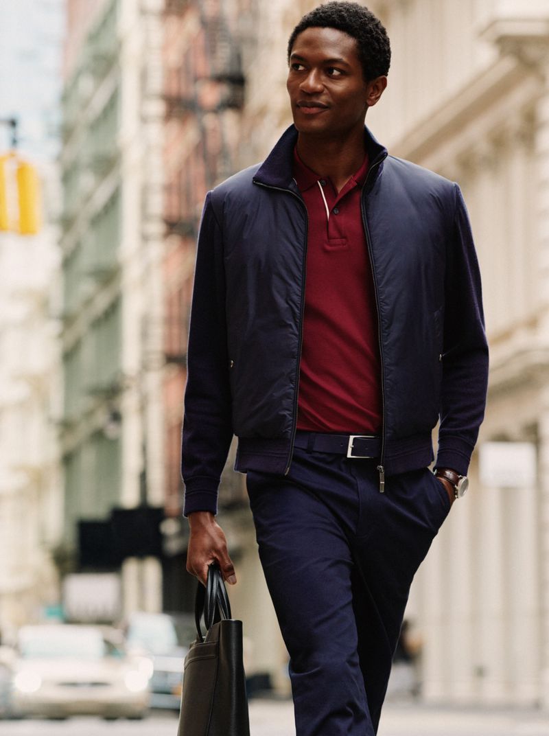 Model Hamid Onifadé in a navy jacket, burgundy polo, and navy trousers holding a black briefcase while walking on a city street.
