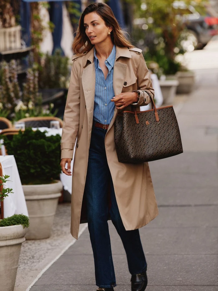 Model Andreea Diaconu walking on a city sidewalk wearing a beige trench coat, blue striped shirt, and holding a monogram tote bag.