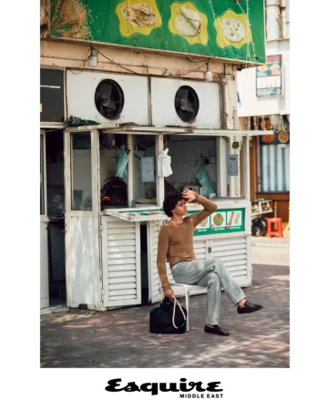 Adult male in casual attire sits on a chair in front of a food stall on a sunny day. The man rests his arm on his forehead.