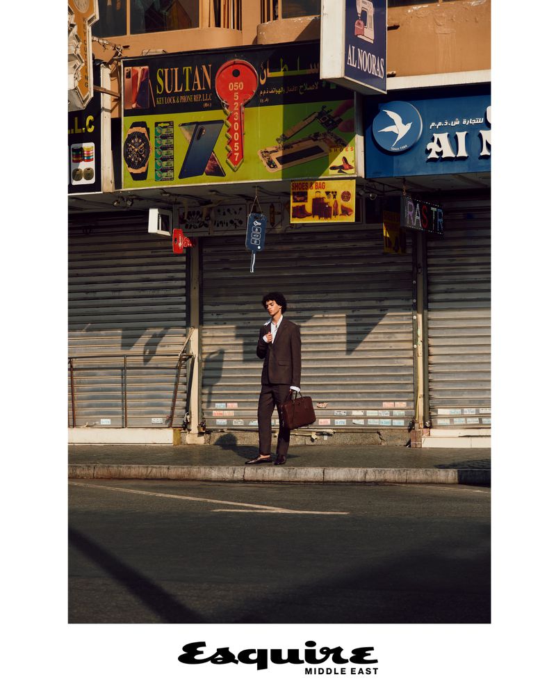 Model Leo Comanescu holding a leather briefcase on a city sidewalk in front of closed shop shutters for Esquire Middle East.