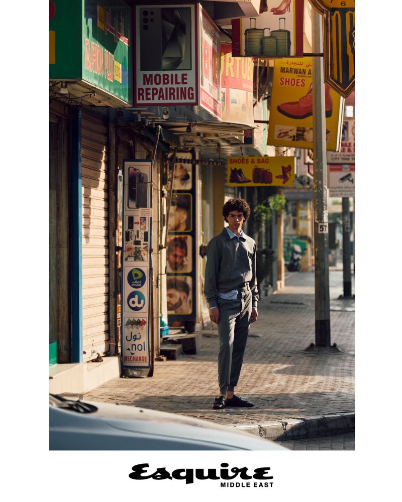 Man in grey suit stands on sunny street in Middle East, posing near store signs. Fashion editorial shot. Esquire Middle East logo shown.