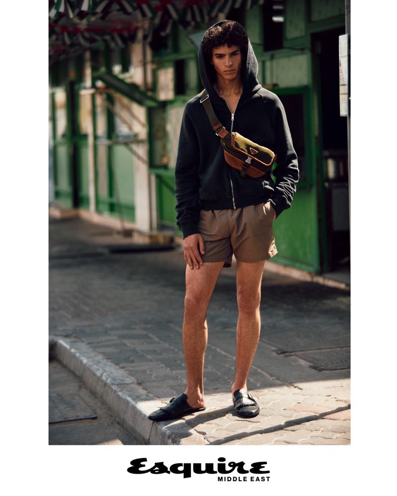 A young adult male poses outdoors wearing a dark zip hoodie, brown shorts, and black loafers, with a brown bag across his chest.