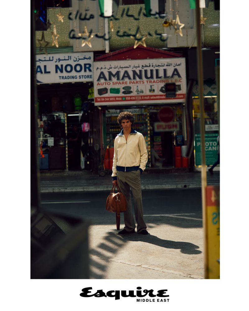 An adult man stands in front of an auto parts store in an urban environment. He is holding a brown leather bag.