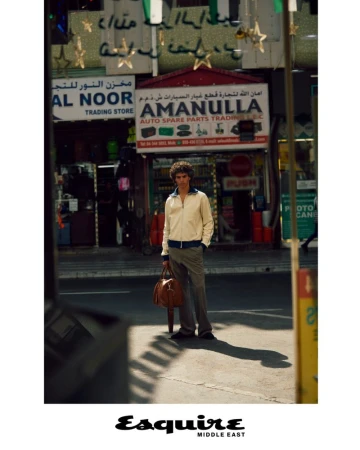 An adult man stands in front of an auto parts store in an urban environment. He is holding a brown leather bag.