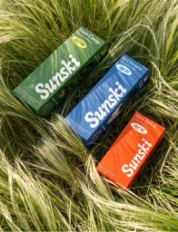 Three colorful Sunski sunscreen boxes in green, blue, and orange arranged on tall ornamental grass in natural sunlight.