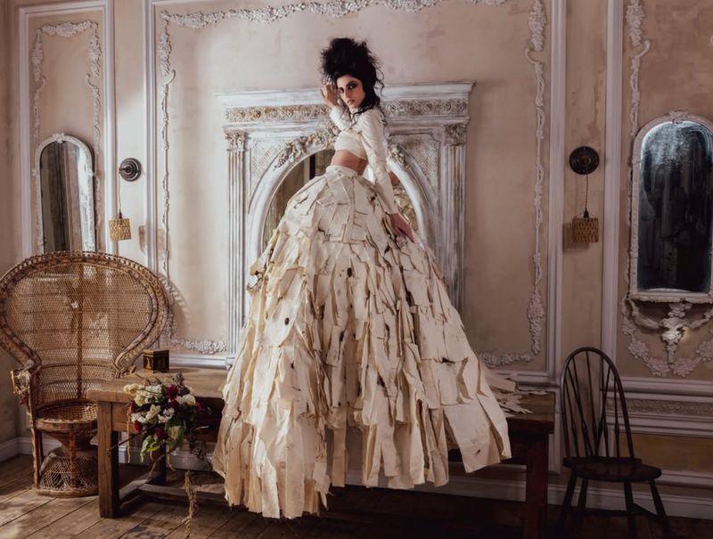 Model wearing a voluminous skirt made of layered paper strips and a long-sleeved cream top, standing inside a classic room with molding.