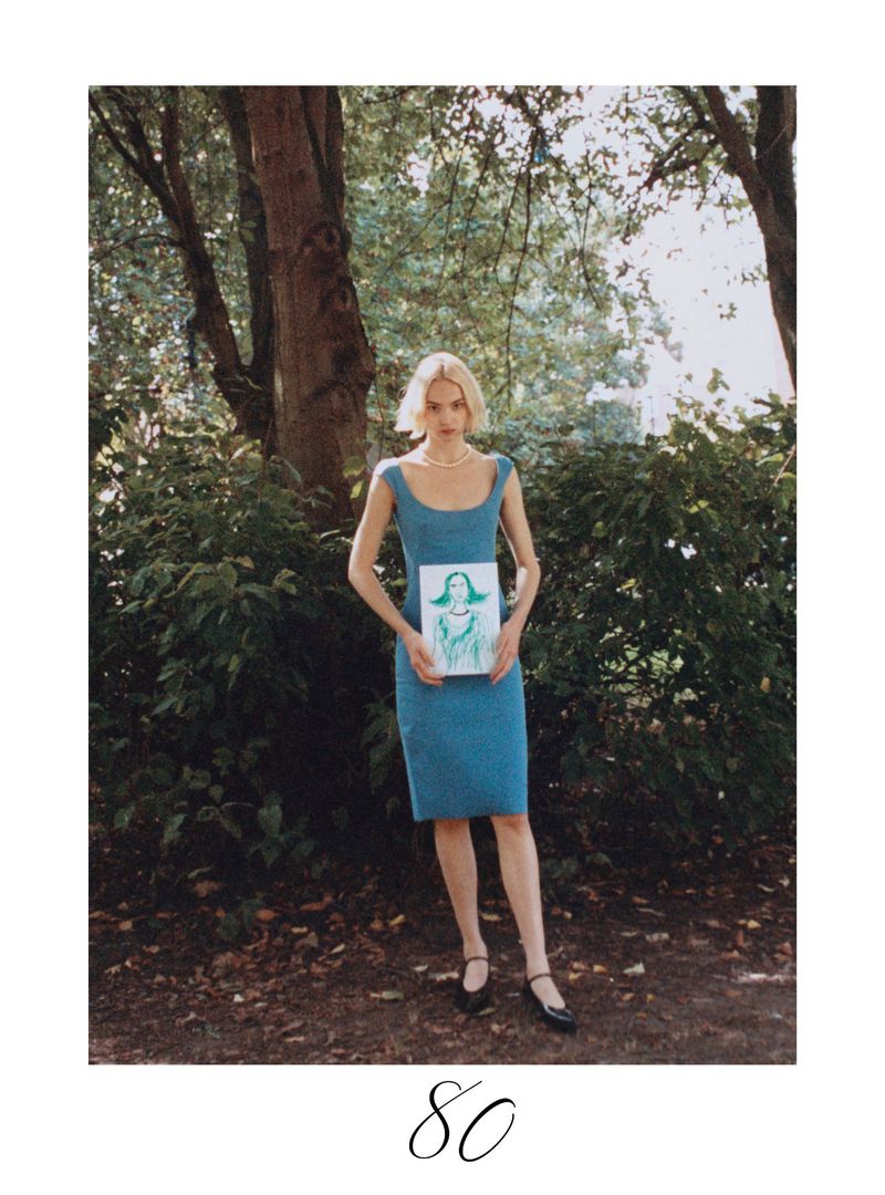 A full shot captures a woman in a blue dress holding a framed illustration in a green, leafy park, illuminated by natural light.