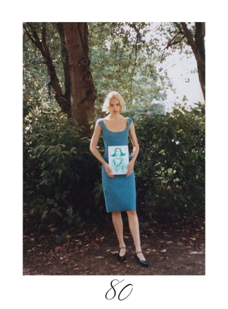 A full shot captures a woman in a blue dress holding a framed illustration in a green, leafy park, illuminated by natural light.