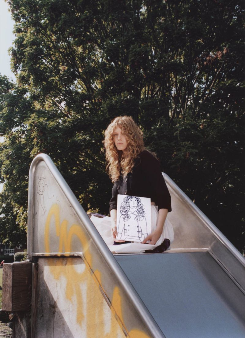 A young adult female sitting on a silver metal playground slide, holding a drawing, with trees and greenery in the background.