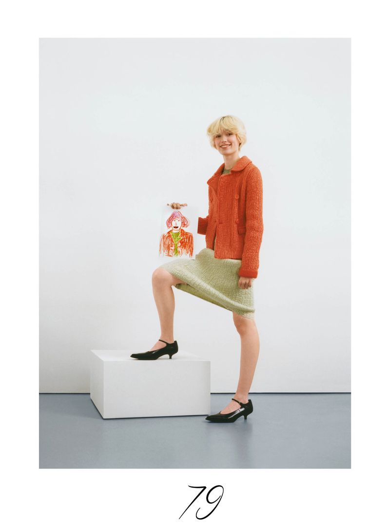 Woman in an orange tweed jacket and light green skirt poses with a handbag that has an illustration, standing on a white box.