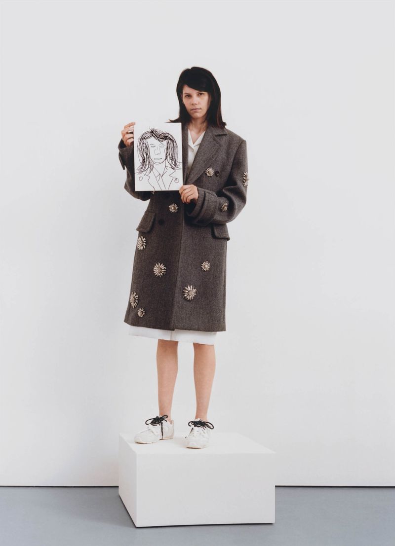Woman in embellished coat stands on white block, holding portrait drawing against a white backdrop in a fashion editorial style photograph.