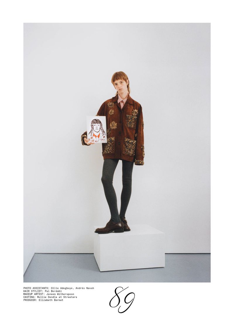 Model standing on a white pedestal in an embellished brown jacket holding a portrait sketch against a white studio background.