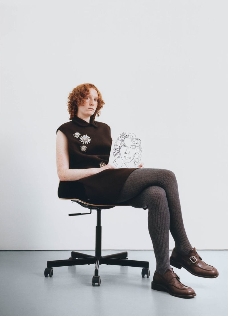 Red-haired adult sitting on a chair holding a sketch of a face in a studio, wearing a brown dress and brown shoes.