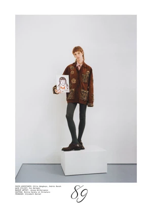 Model standing on a white pedestal in an embellished brown jacket holding a portrait sketch against a white studio background.