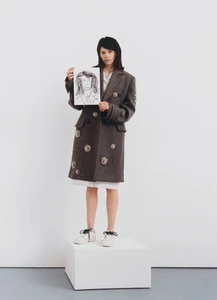 Woman standing on a white cube holding a framed portrait illustration wearing a grey embellished coat and white sneakers against a white wall