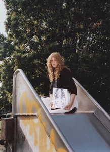 Model with wavy curly hair sits atop a metal playground slide holding a line-art portrait illustration in an outdoor park setting.