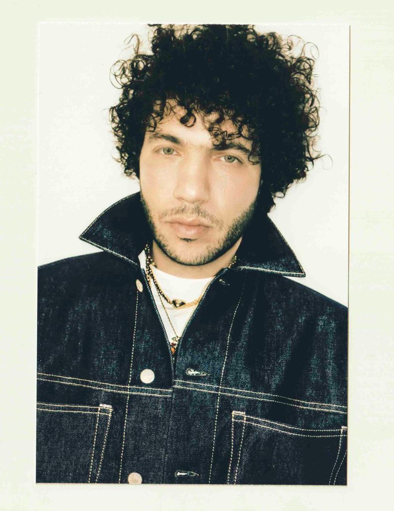 Benny Blanco wearing a dark denim jacket with gold necklaces against a plain white background.