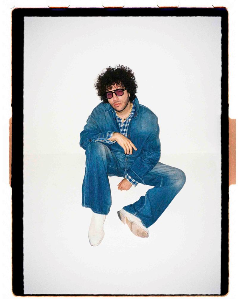 Benny Blanco wearing a denim jacket, matching jeans, and tinted sunglasses while seated against a white background.