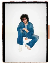 Benny Blanco wearing a denim jacket, matching jeans, and tinted sunglasses while seated against a white background.