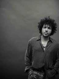 Benny Blanco in a striped button-up shirt and chain necklace posing against a dark grey studio backdrop in black and white.