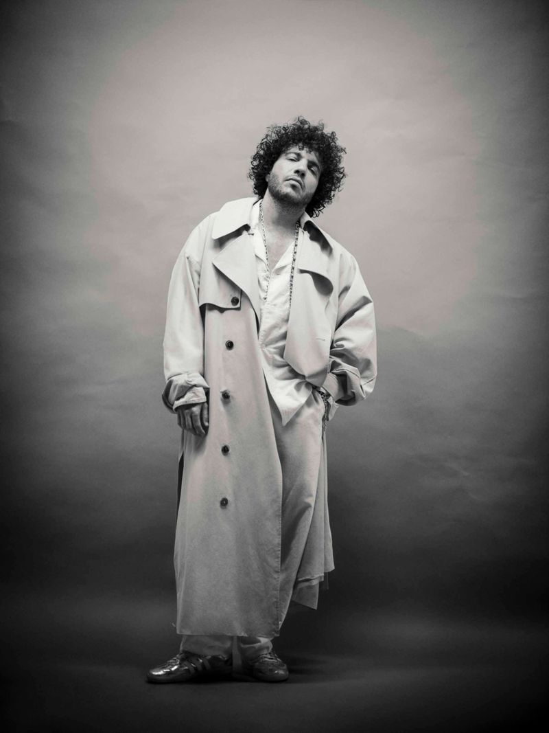 Benny Blanco standing in an oversized light-colored trench coat and loose trousers against a studio backdrop in black and white.