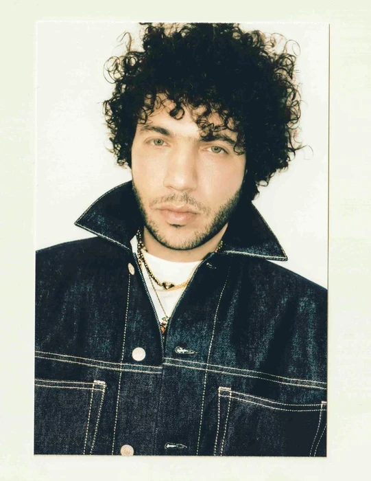 Benny Blanco wearing a dark denim jacket with gold necklaces against a plain white background.