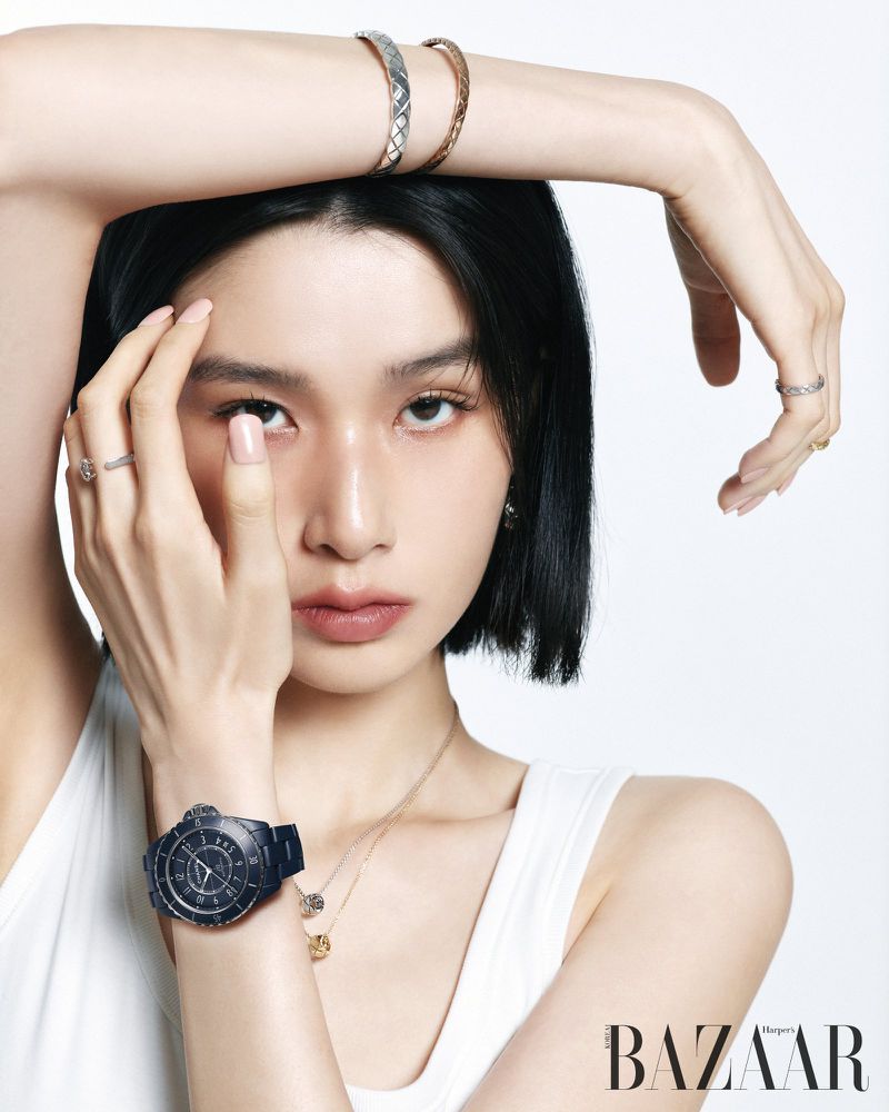 Esther Kim posing with arms raised wearing a dark blue luxury watch, silver bangles, and rings against a white backdrop.