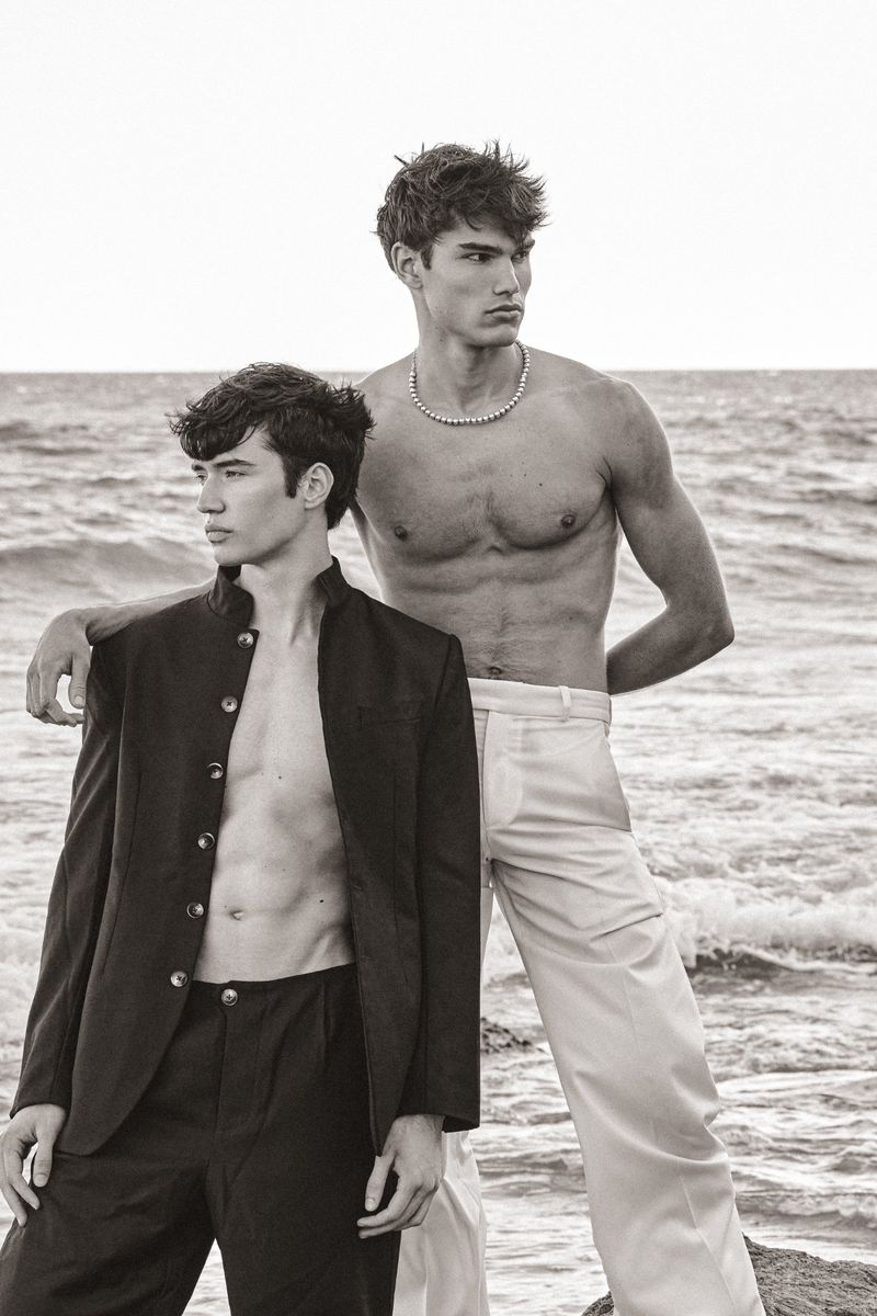 Two men standing on a beach; one wears a black open jacket and trousers, the other is shirtless with white trousers and a bead necklace.