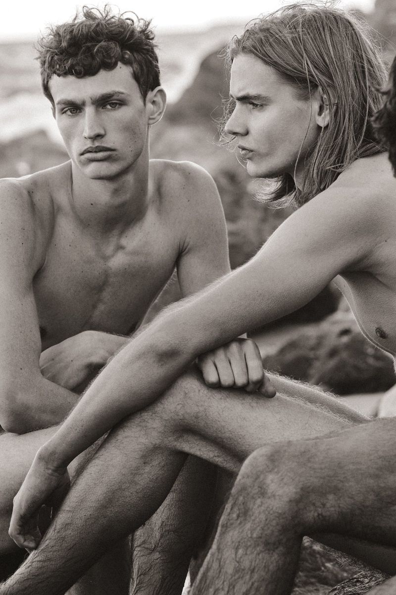 Black and white editorial photograph of shirtless young men seated together along a rocky coastal shoreline.