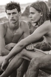 Black and white editorial photograph of shirtless young men seated together along a rocky coastal shoreline.