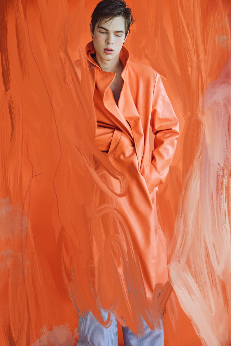 Young man with light skin wearing a vibrant orange trench coat, partially obscured by painted orange brushstrokes on a studio background.