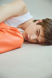 Young model lying down on a light studio surface wearing a white ribbed tank top and bright peach trousers.
